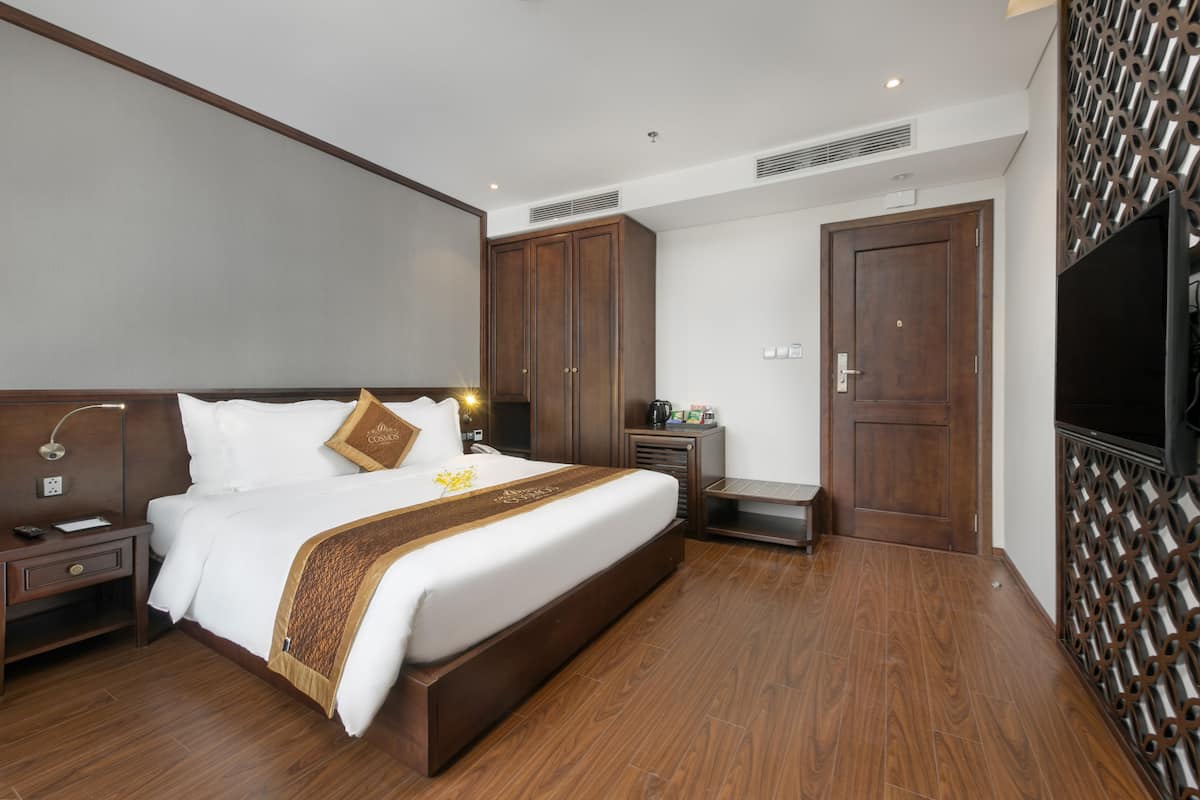 A hotel room with a large bed, wooden headboard, a TV, a desk, and a decorative wall panel.