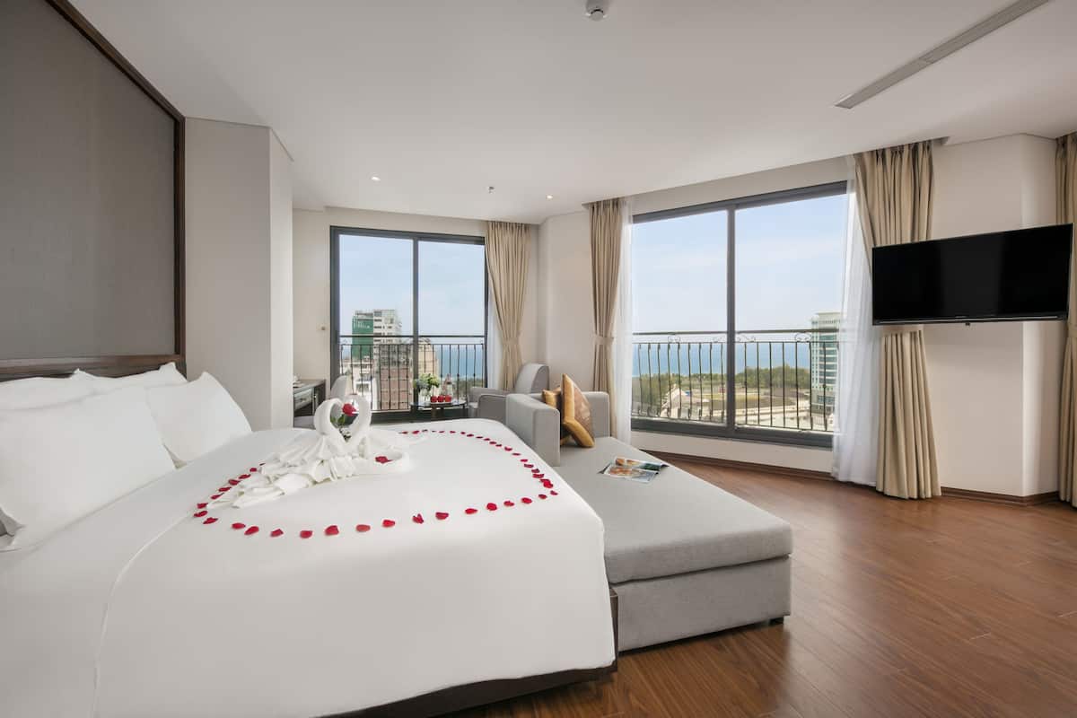A modern hotel room with a large bed, a grey sofa, a flat-screen TV, and a balcony with a view of the city.