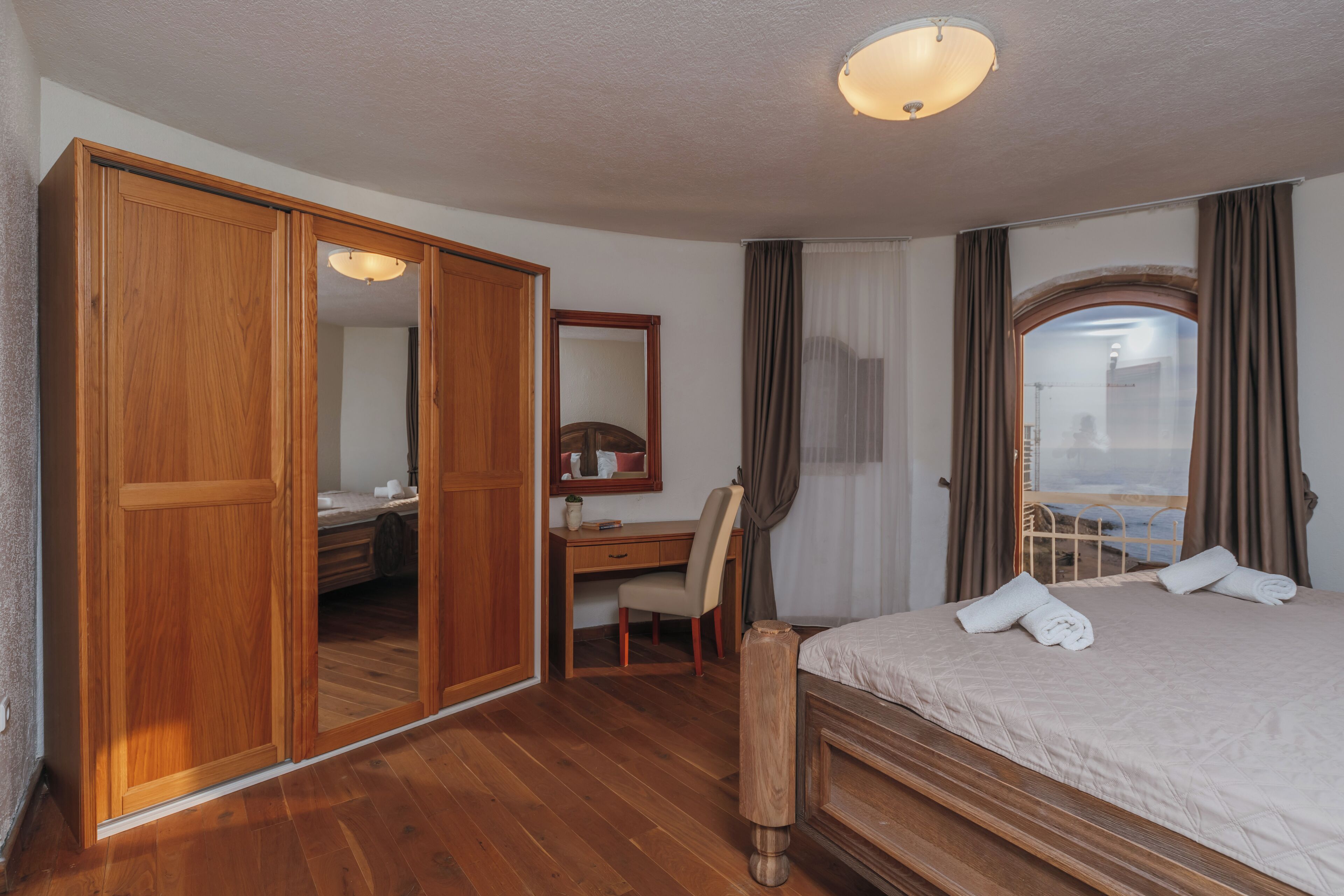 A hotel room with a wooden wardrobe, a desk with a chair, a bed with towels, and a large mirror.