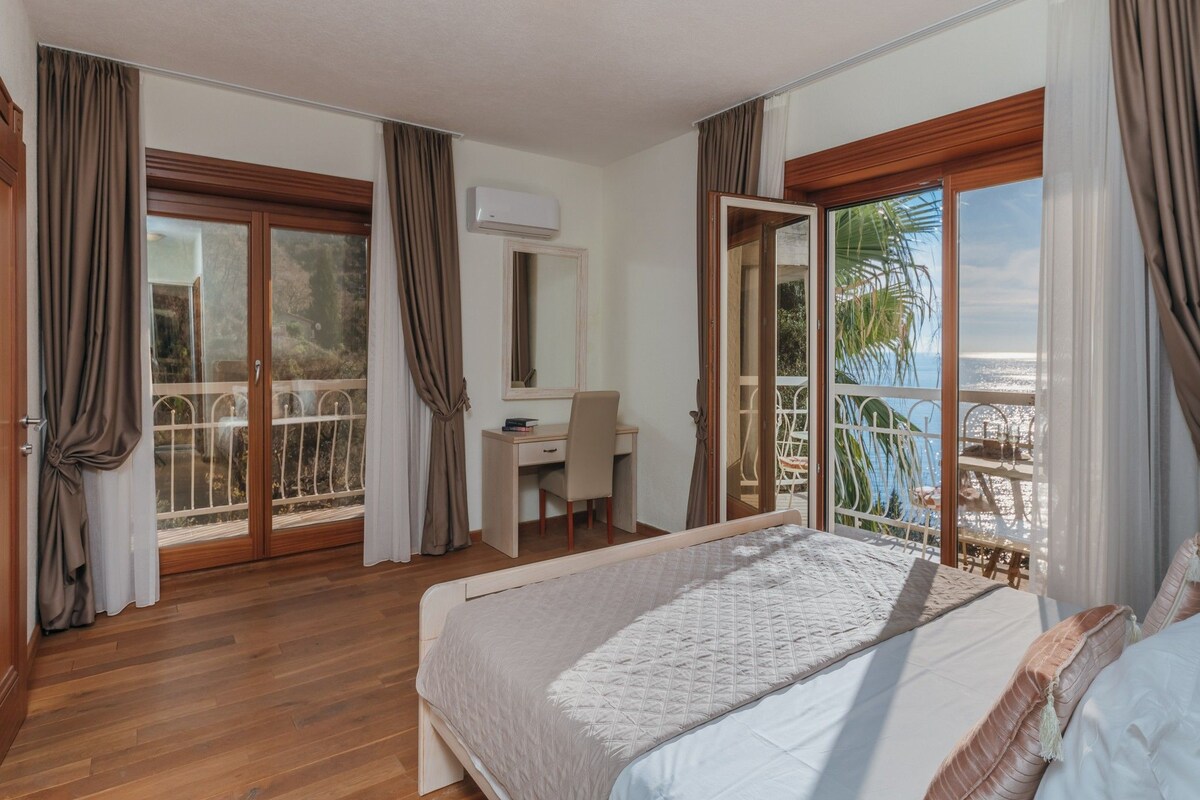 A bedroom with a bed, a desk, and a chair. There is a balcony with a view of the sea.