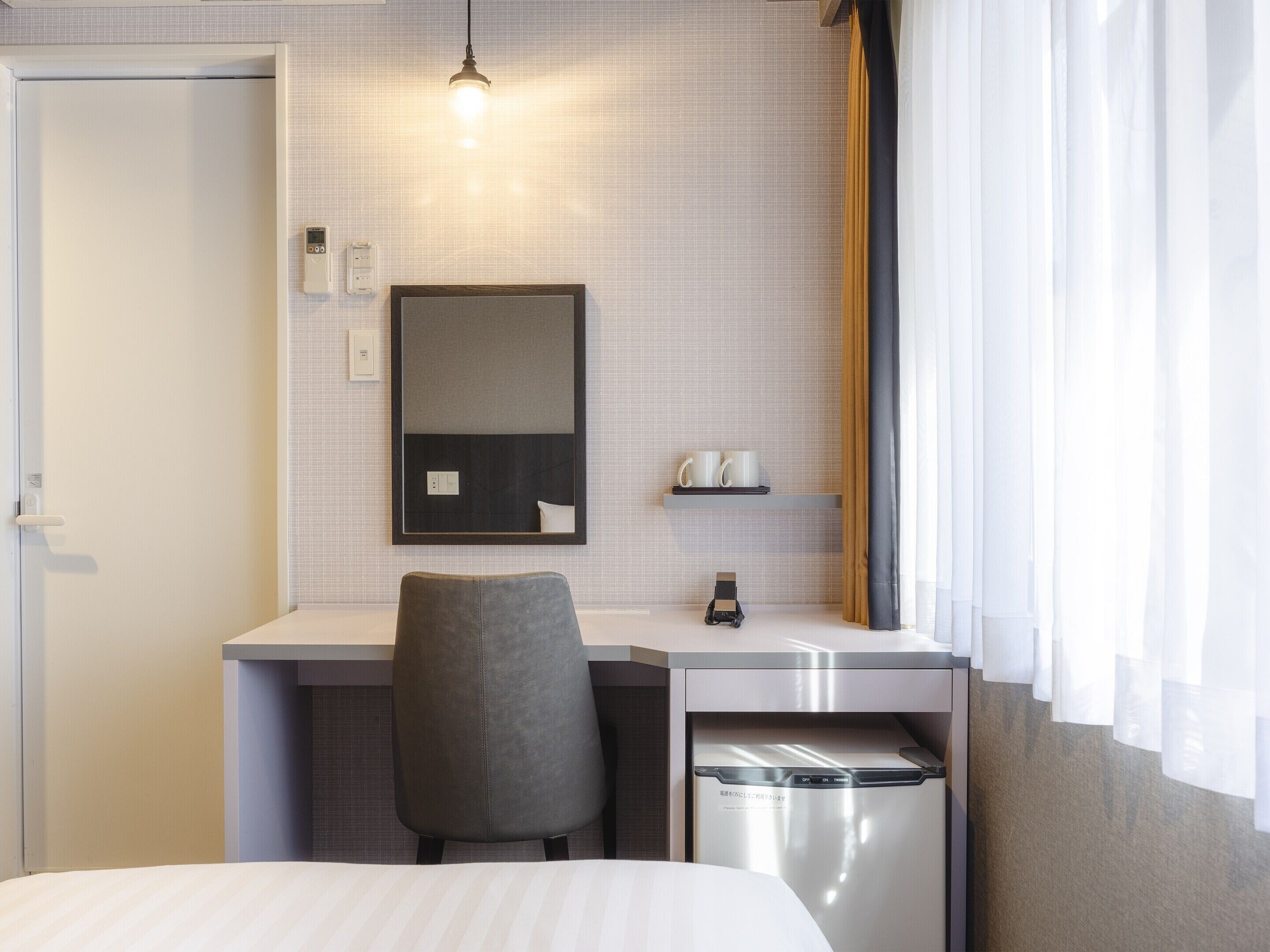 Una habitación de hotel con una cama, un escritorio con una silla, un espejo, un estante con dos tazas, un cubo de basura pequeño y una ventana con cortinas.