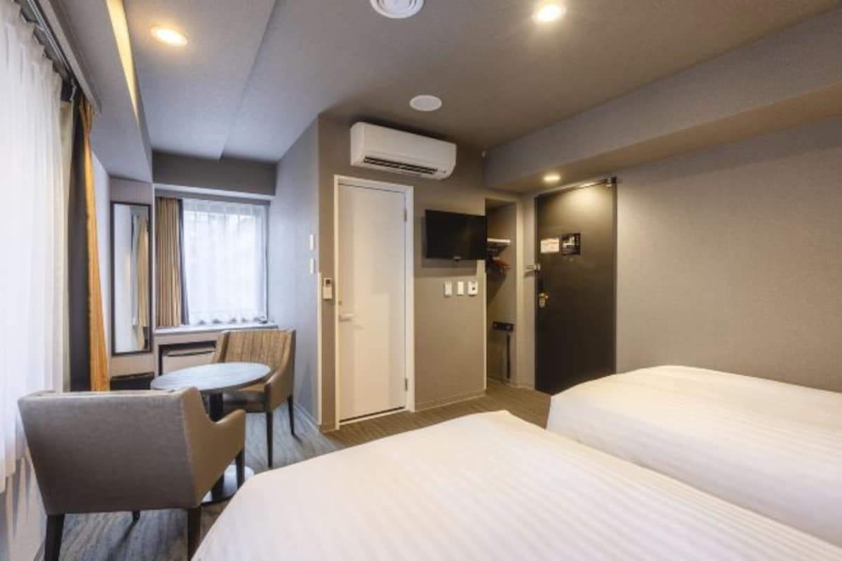 A hotel room with two beds, a dining table, a chair, an air conditioning unit, a television, and a door leading to a bathroom.