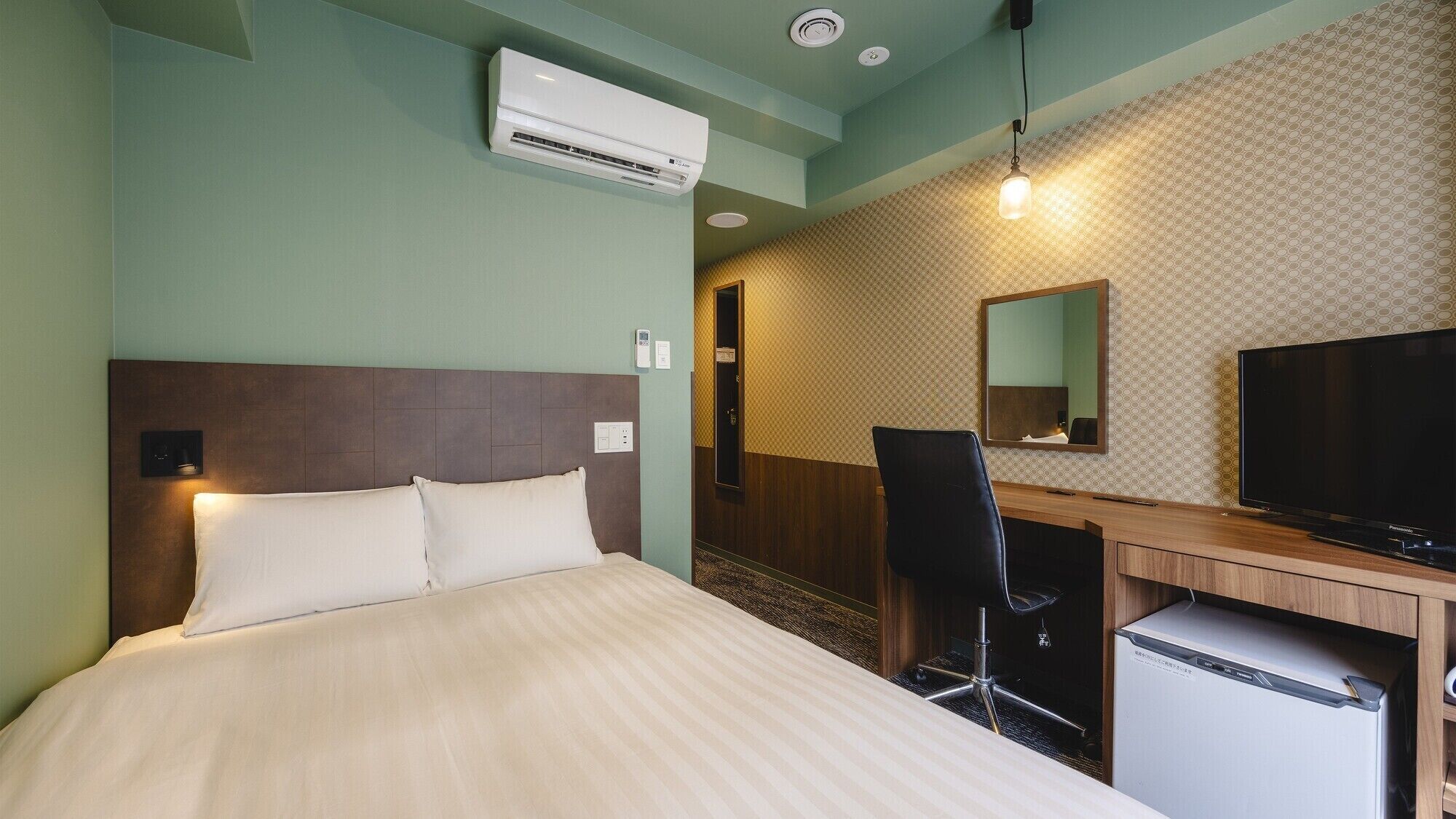A hotel room with a bed, desk, chair, TV, air conditioning unit, and a mirror.