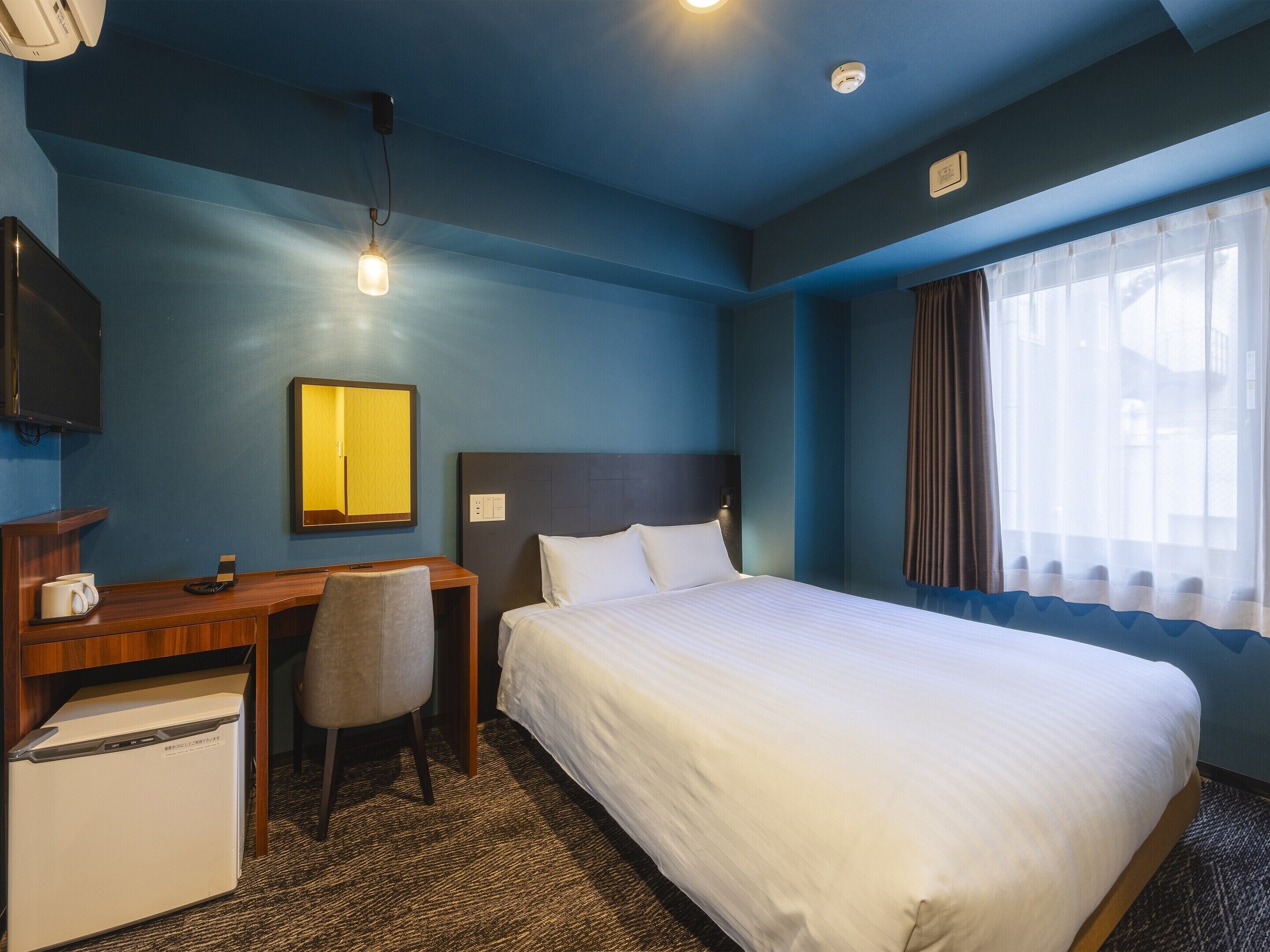 Una habitación de hotel con cama, escritorio, silla, televisor y ventana con cortinas.