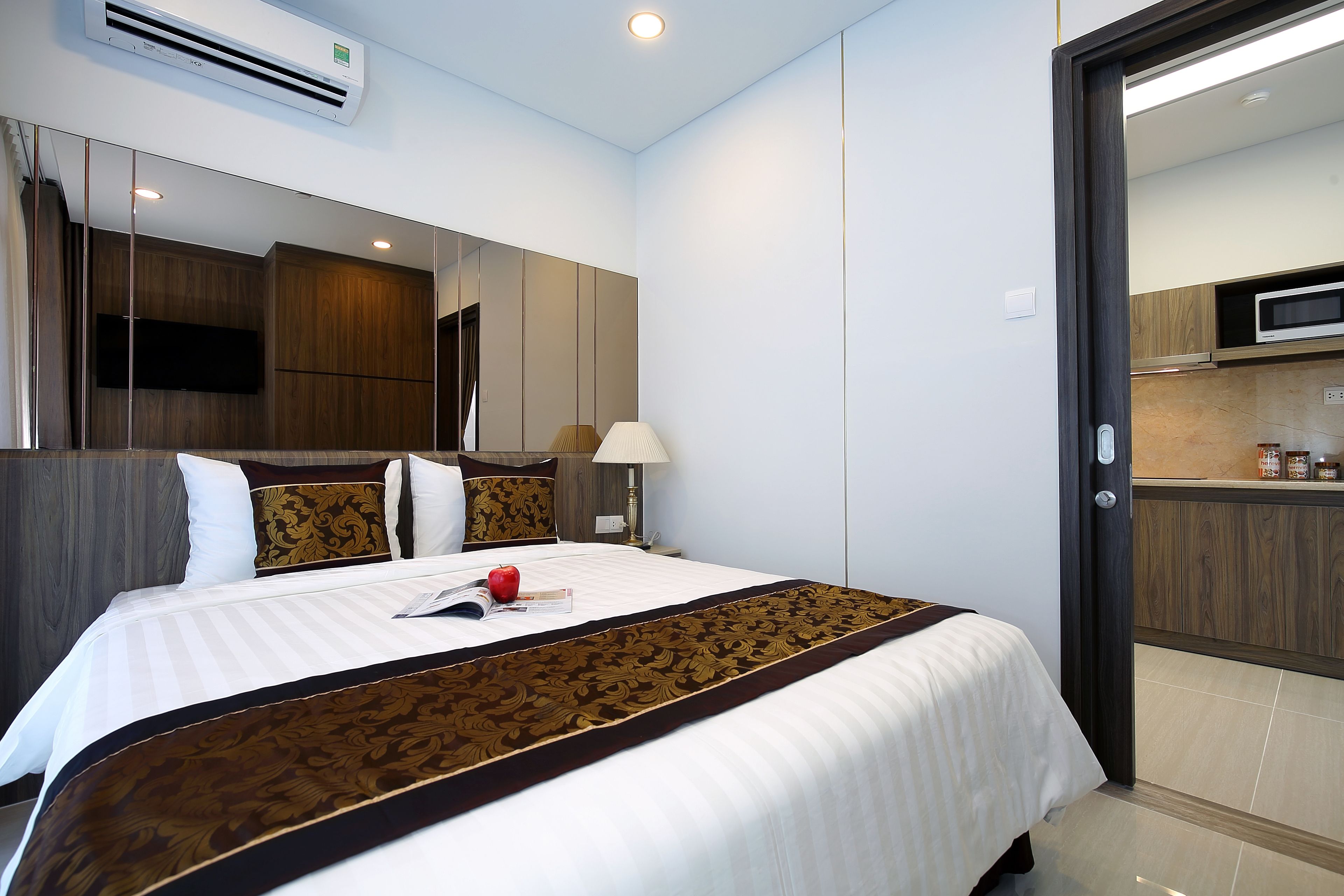 A hotel room with a large bed, a mirror, a bedside lamp, and a view of the kitchen through an open door.