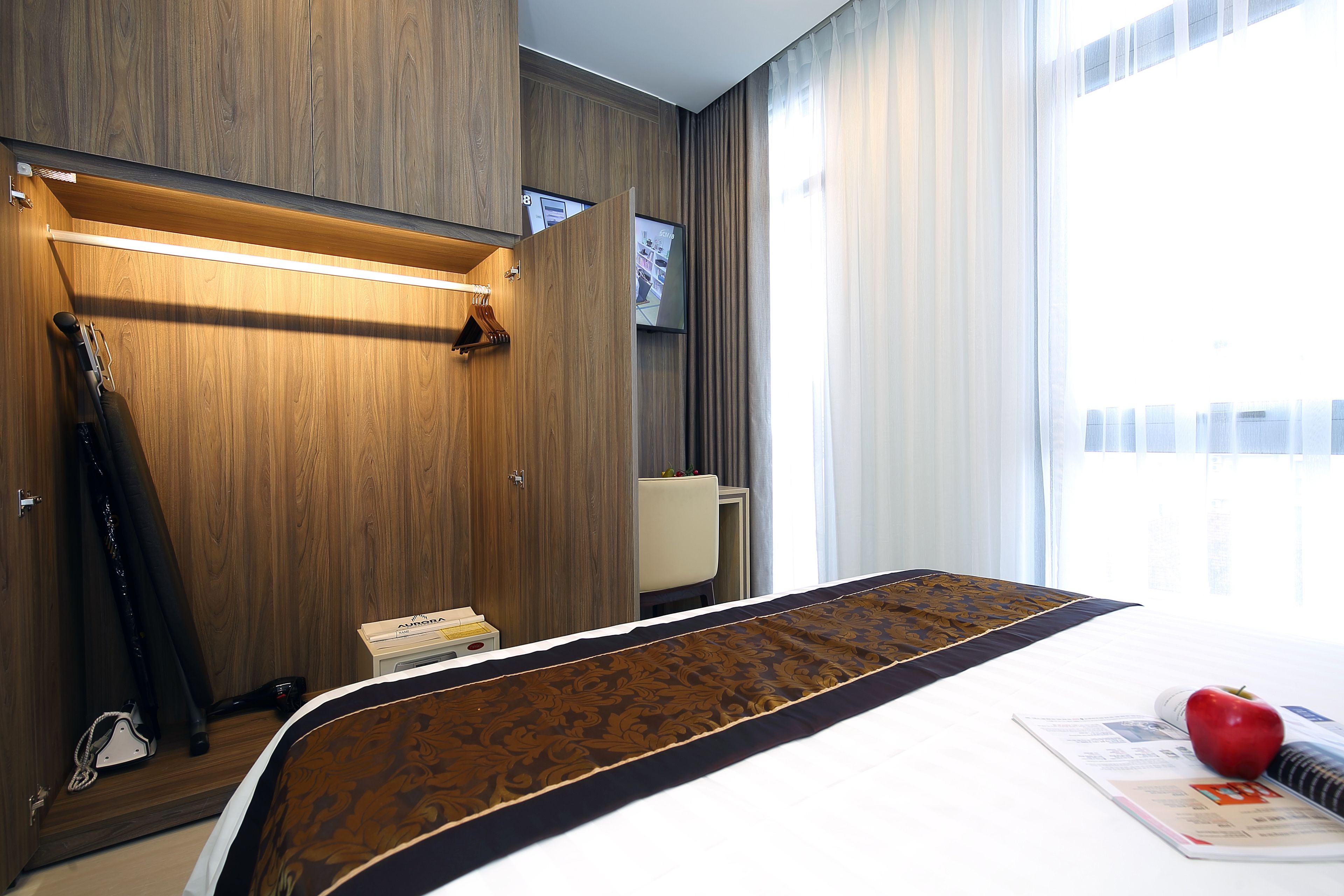 Une chambre d’hôtel avec une armoire en bois, un lit recouvert d’une courtepointe à motifs, une pomme rouge posée sur le lit et une fenêtre avec des rideaux.