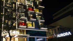 Un hôtel moderne à plusieurs étages, doté de balcons illuminés, d’une entrée bien visible et d’une enseigne portant le nom « AURORA ».