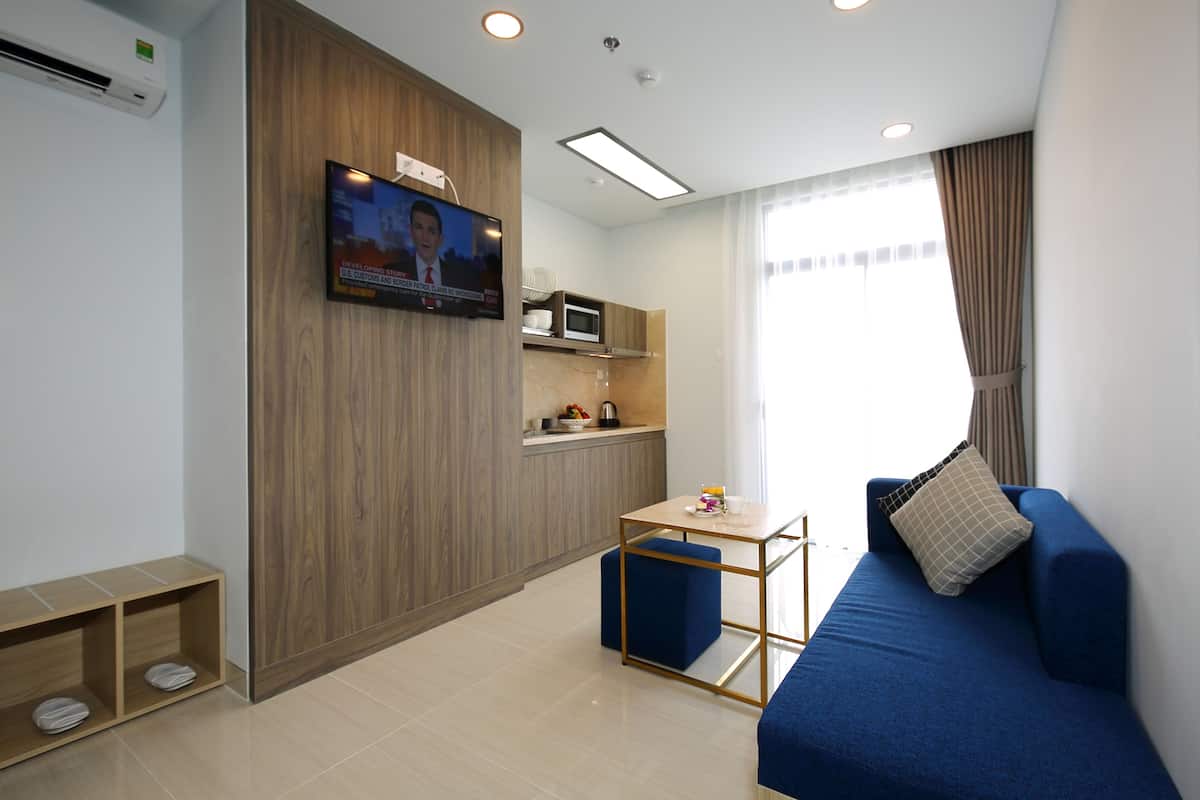 Ein modernes Hotelzimmer mit an der Wand montiertem Fernseher, einer Küchenzeile, einem blauen Sofa und einem kleinen Tisch mit Kissen.