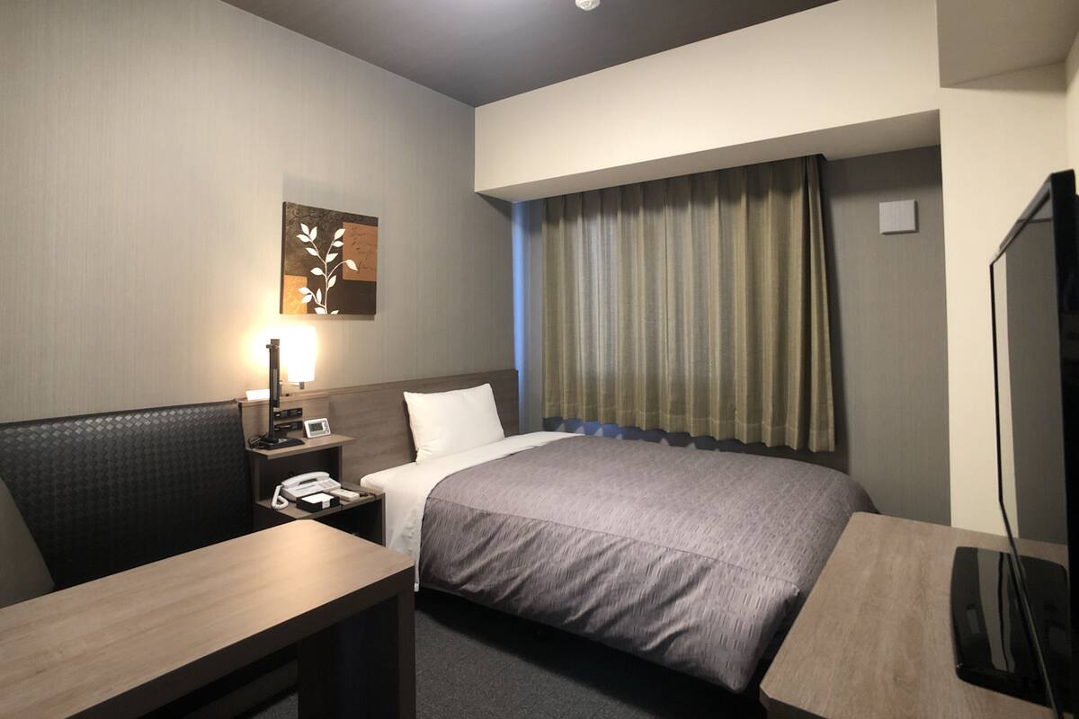Une chambre d’hôtel comprenant un lit, un bureau, une table de chevet, une lampe, un téléviseur et une fenêtre avec des rideaux.