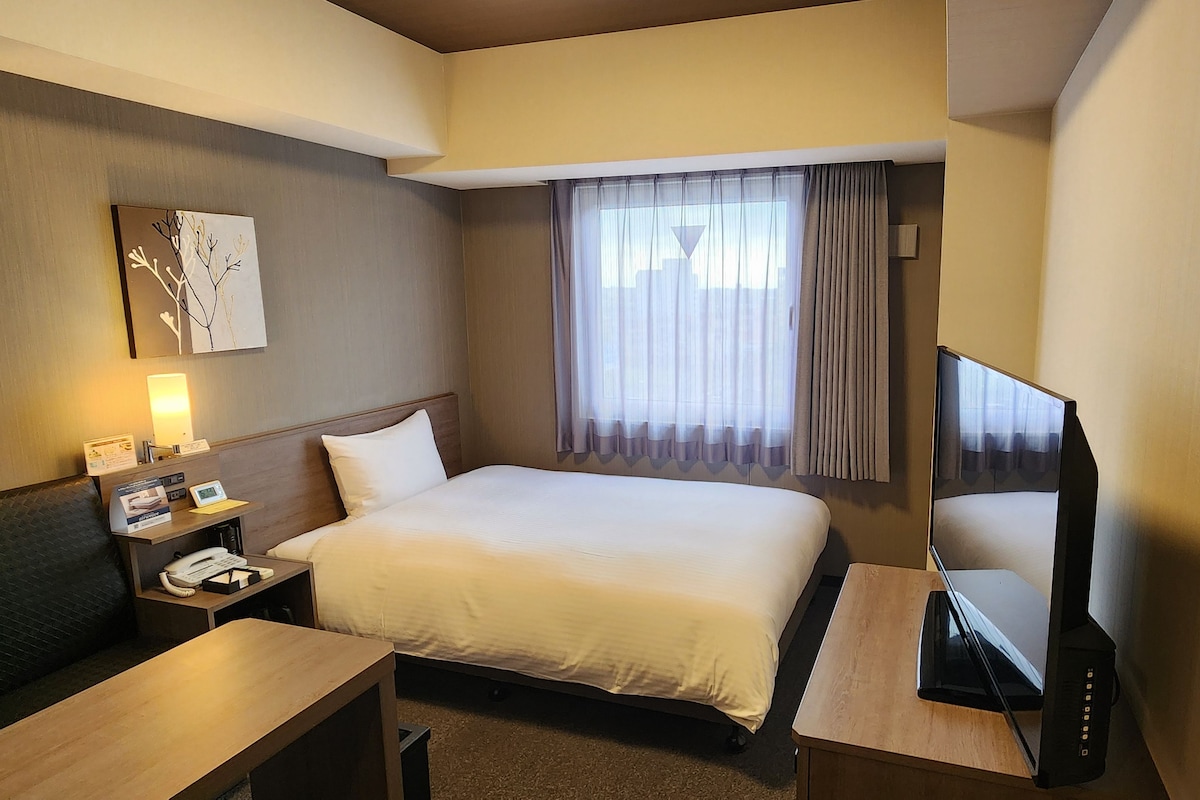 Une chambre d’hôtel avec un lit, un bureau avec un téléphone, une télévision et une fenêtre avec des rideaux.