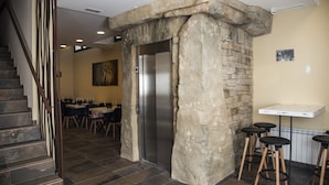 Entrada de elevador com paredes de pedra, área de refeições com mesas e cadeiras, e um bar com banquetas.