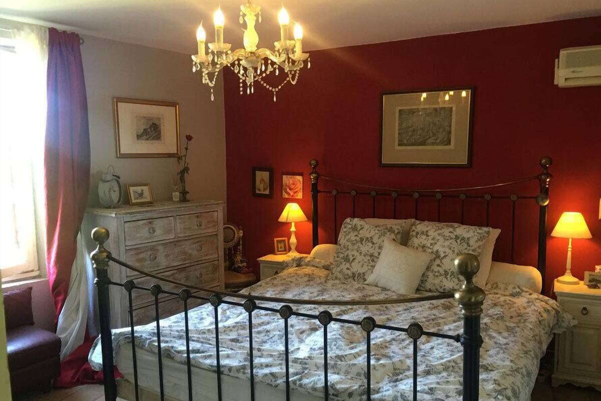 A bedroom with a four-poster bed, a chandelier, a dresser, and a window with curtains.