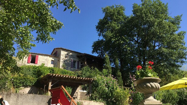 A rustic stone house with a red roof, surrounded by lush greenery and a garden with a stone fountain and a yellow umbrella. - Le Peyrail (Les Lèves-et-Thoumeyragues)