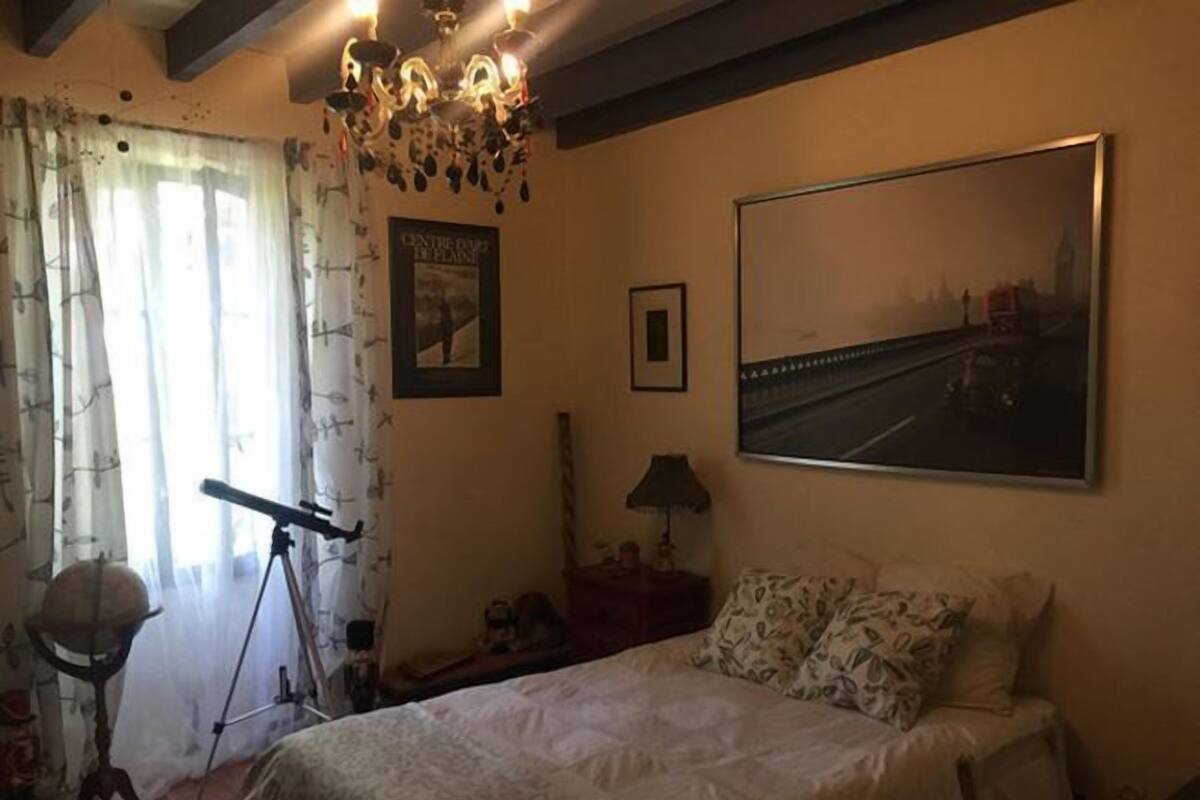 A bedroom with a bed, a large window with curtains, a chandelier, and a framed picture on the wall.