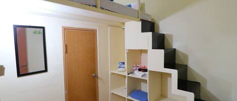 Una habitación con una cama individual alta y otra debajo, una puerta, un espejo y una pequeña estantería con varios objetos.