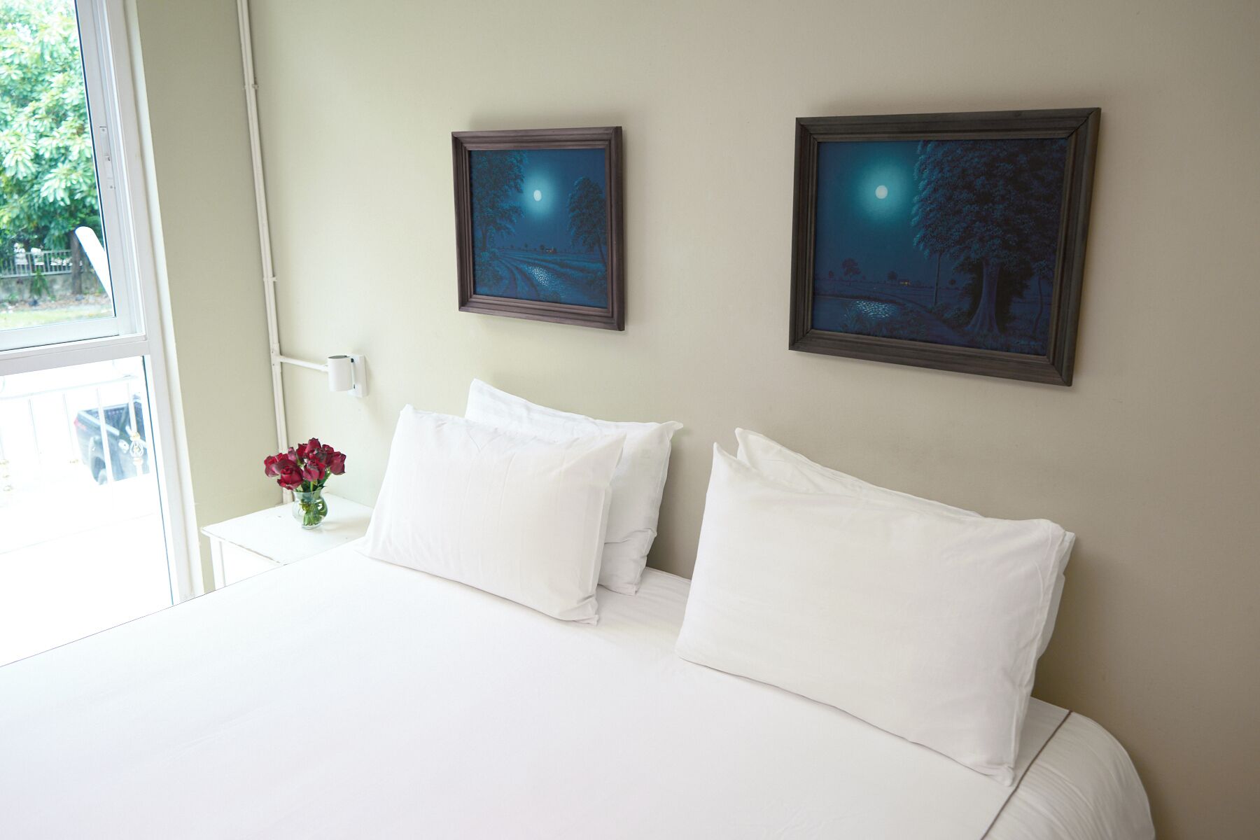 A hotel room with a bed, white pillows, a vase with red flowers, and two framed pictures on the wall.