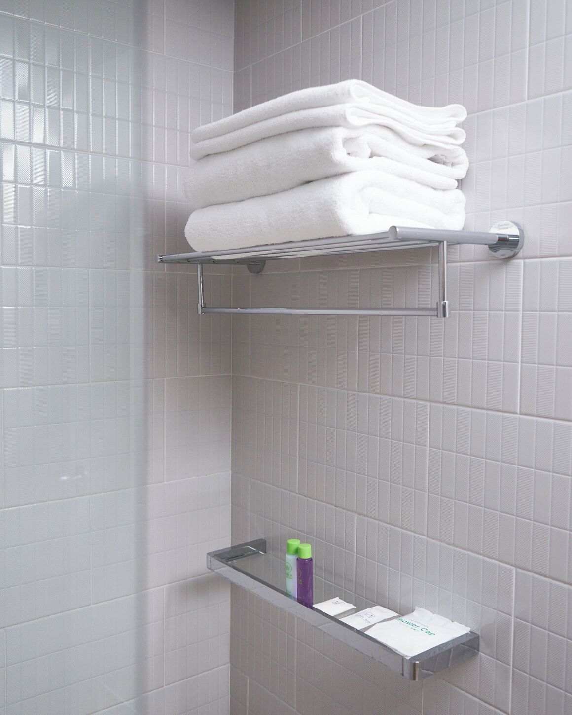 Un baño con paredes de azulejos blancos, un perchero con toallas dobladas y un estante con artículos de aseo.