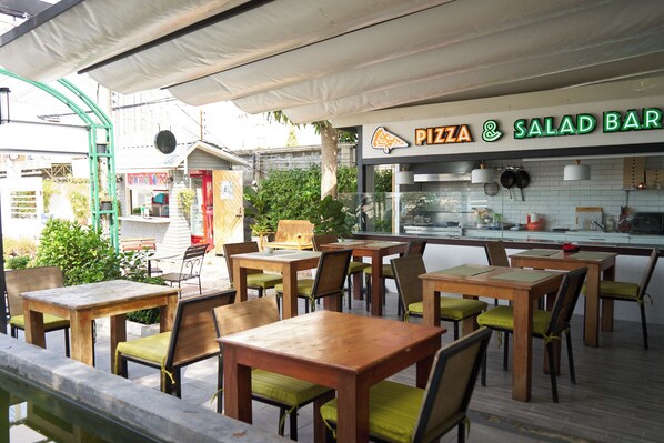Un espacio exterior con mesas y sillas, una cocina visible a través del vidrio, y un letrero para una barra de pizza y ensaladas.