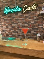 Una zona de bar con un letrero de neón que dice "Warila Café", una copa de martini con una bebida roja, un coctelera y una botella de ginebra sobre la barra.
