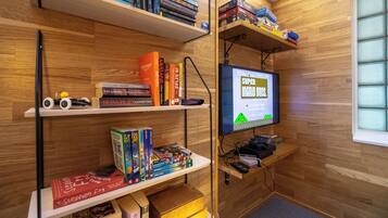 Una habitación con paredes de madera, estantes con libros y juegos, un televisor mostrando un juego y una ventana.