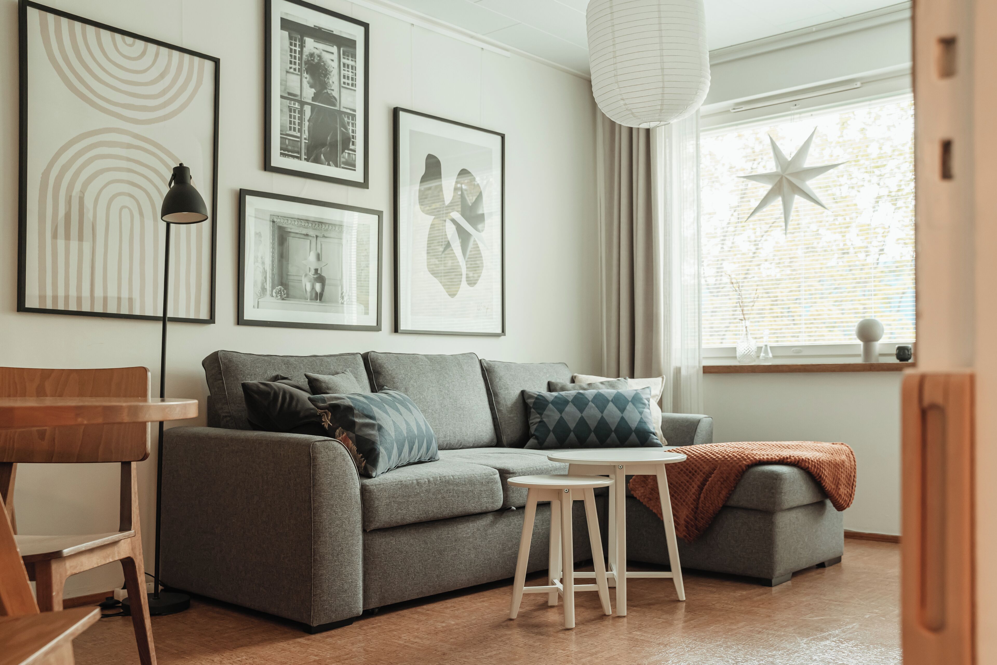Una sala de estar con un sofá gris, una mesita de madera y una lámpara de pie. Se observan cuadros enmarcados en la pared y una decoración con forma de estrella en el alféizar de la ventana.
