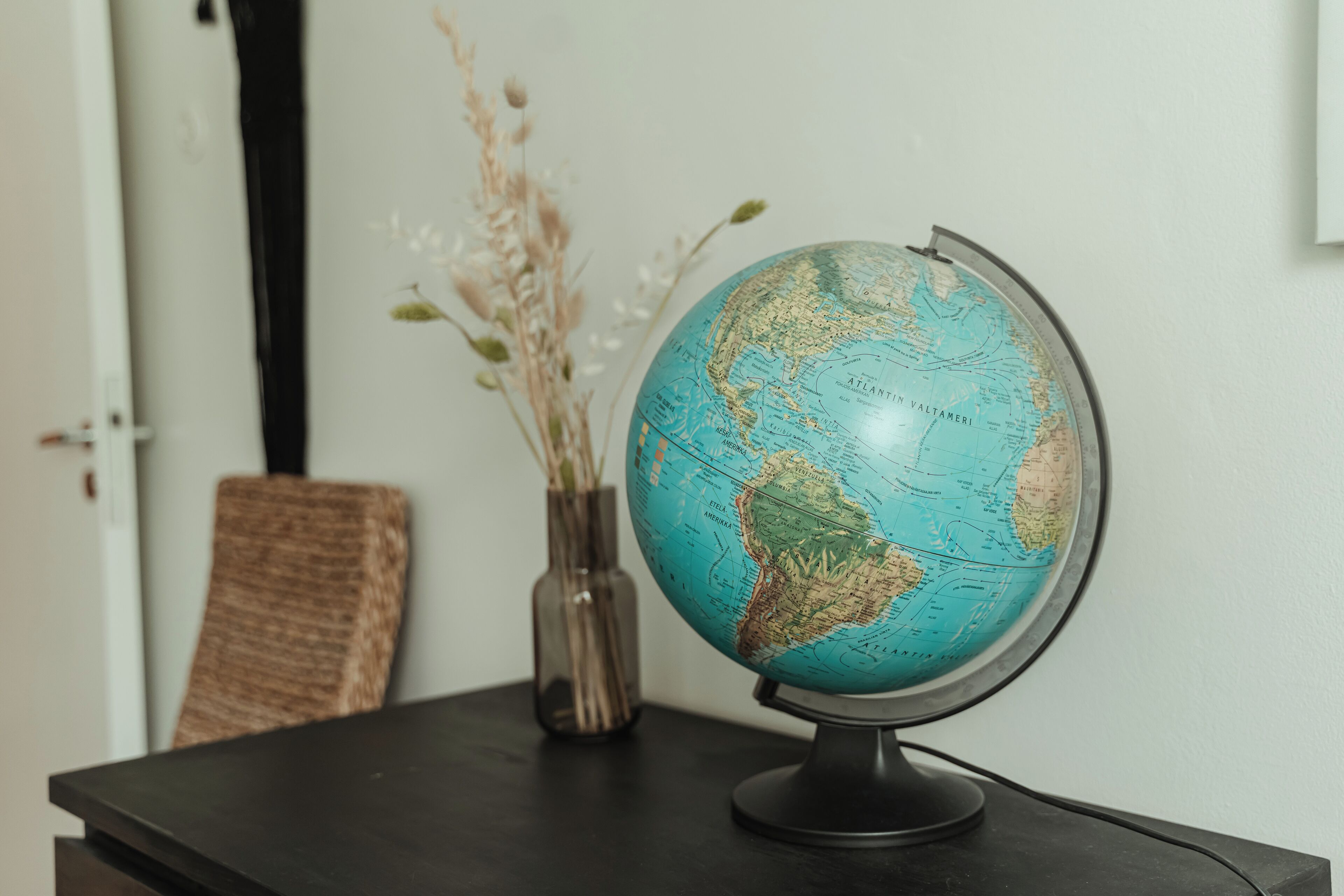 A globe on a stand on a table, with a vase of dried flowers and a chair in the background.