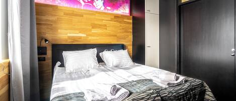 A modern hotel room with a wooden headboard, a large bed with white linens, and a digital display above the bed.