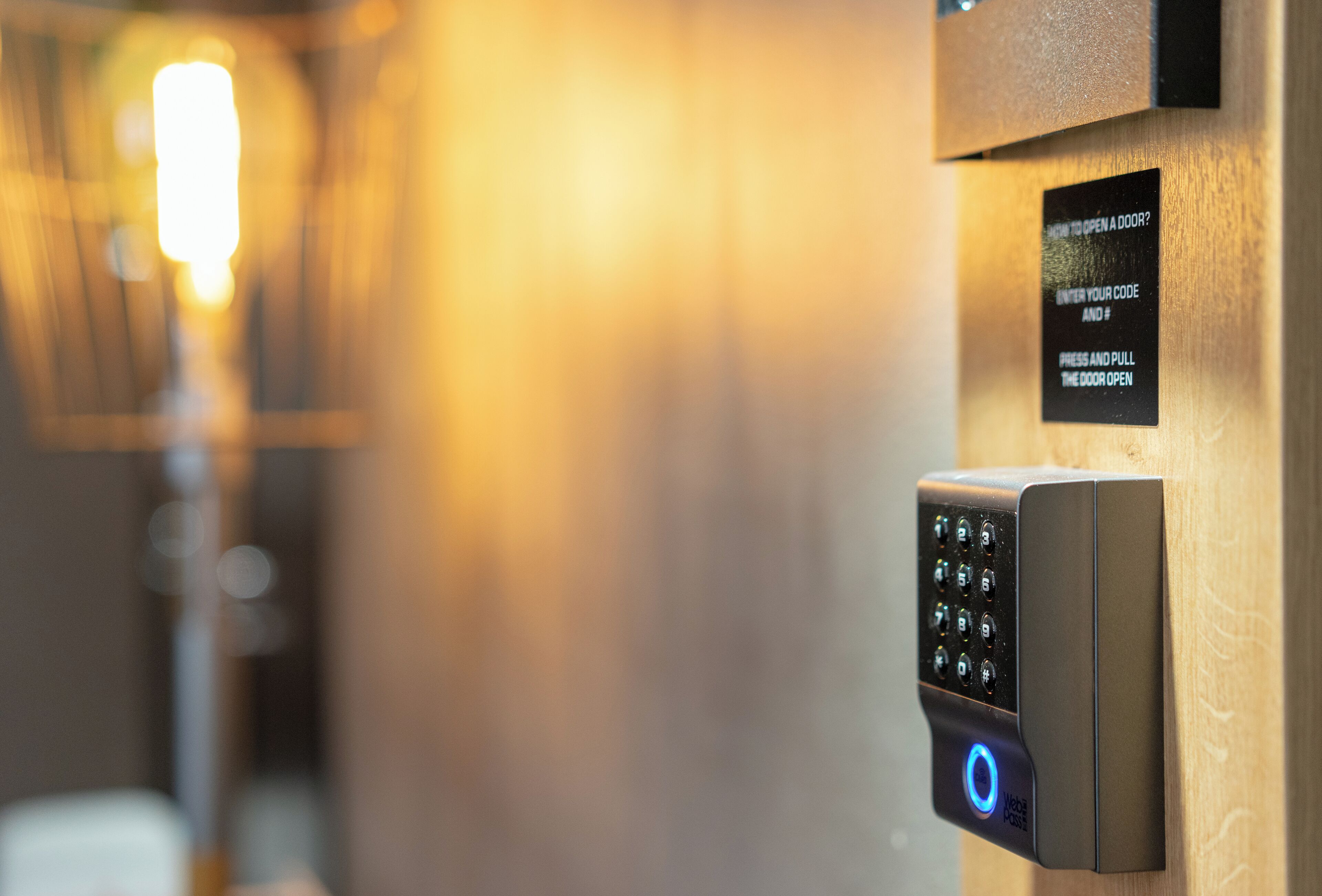 A modern door lock with a keypad and a digital display, a sign instructing to enter a code and pull the door open, and a blurred background suggesting an indoor setting.
