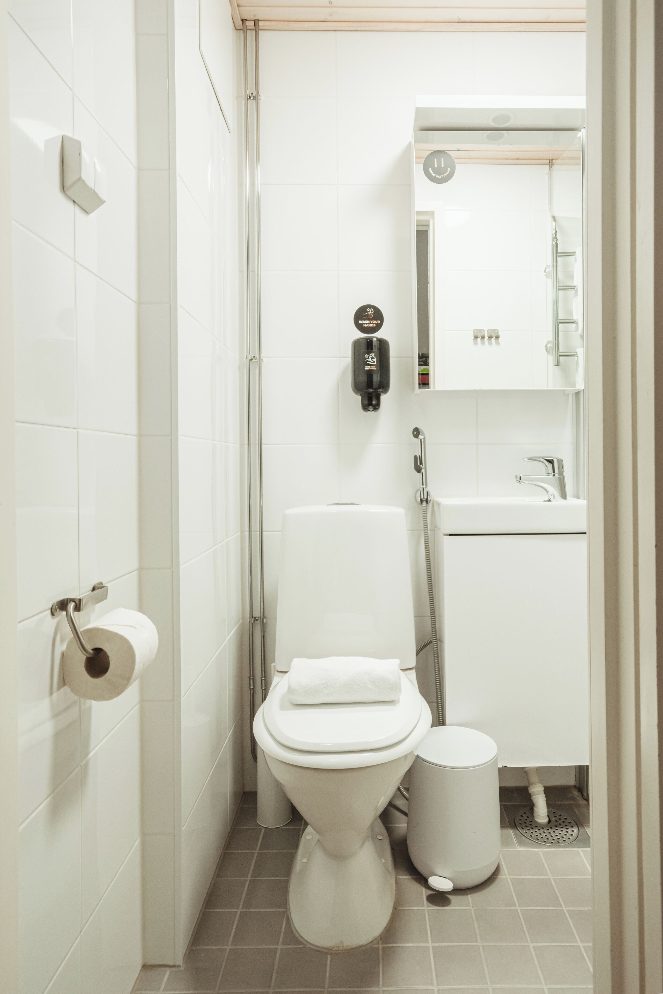 Un baño pequeño y limpio con inodoro, un bote de basura, un secador de manos y un espejo sobre el lavamanos.