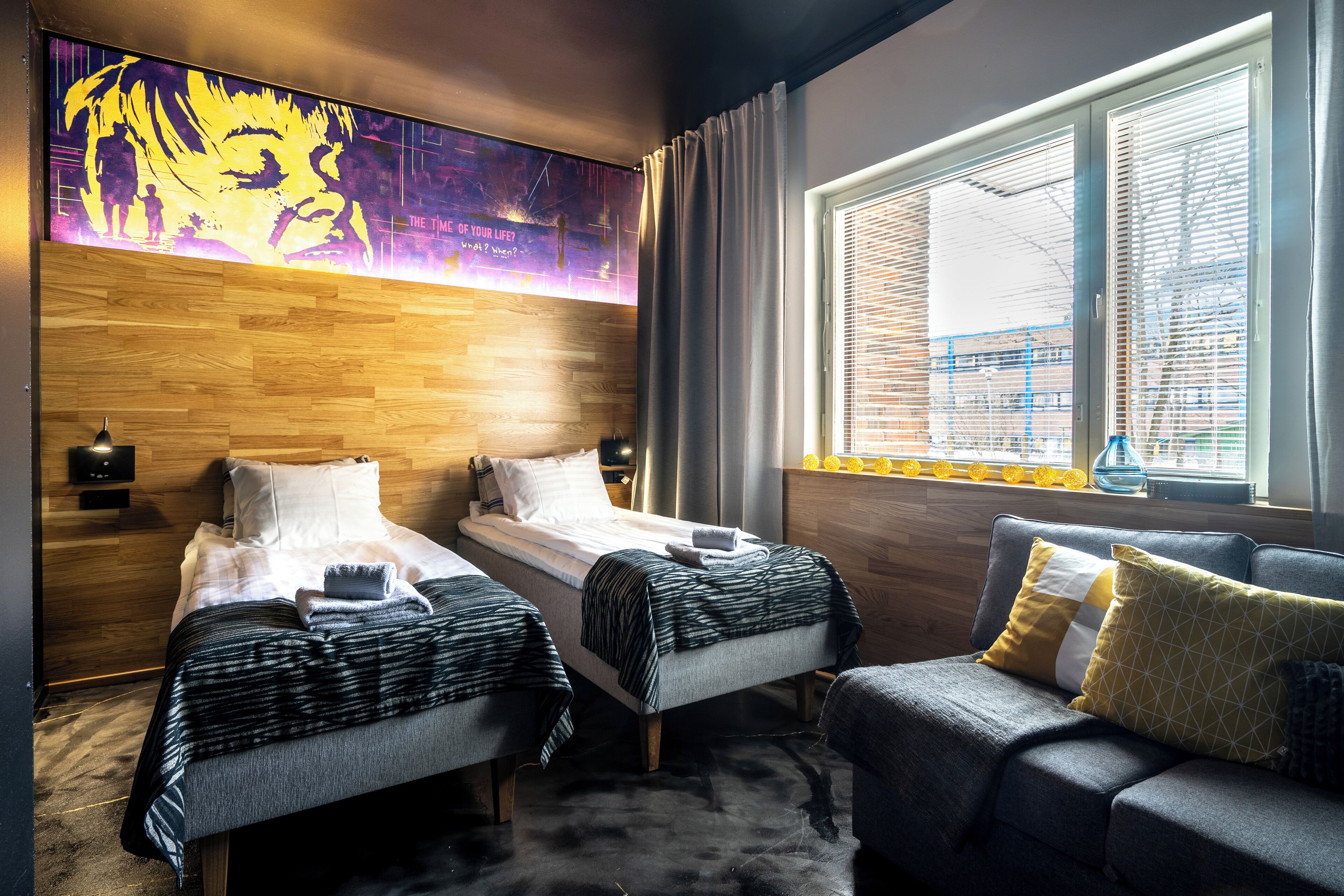 A hotel room with two single beds, a sofa, a large window with blinds, a wooden headboard, and a wall mural.