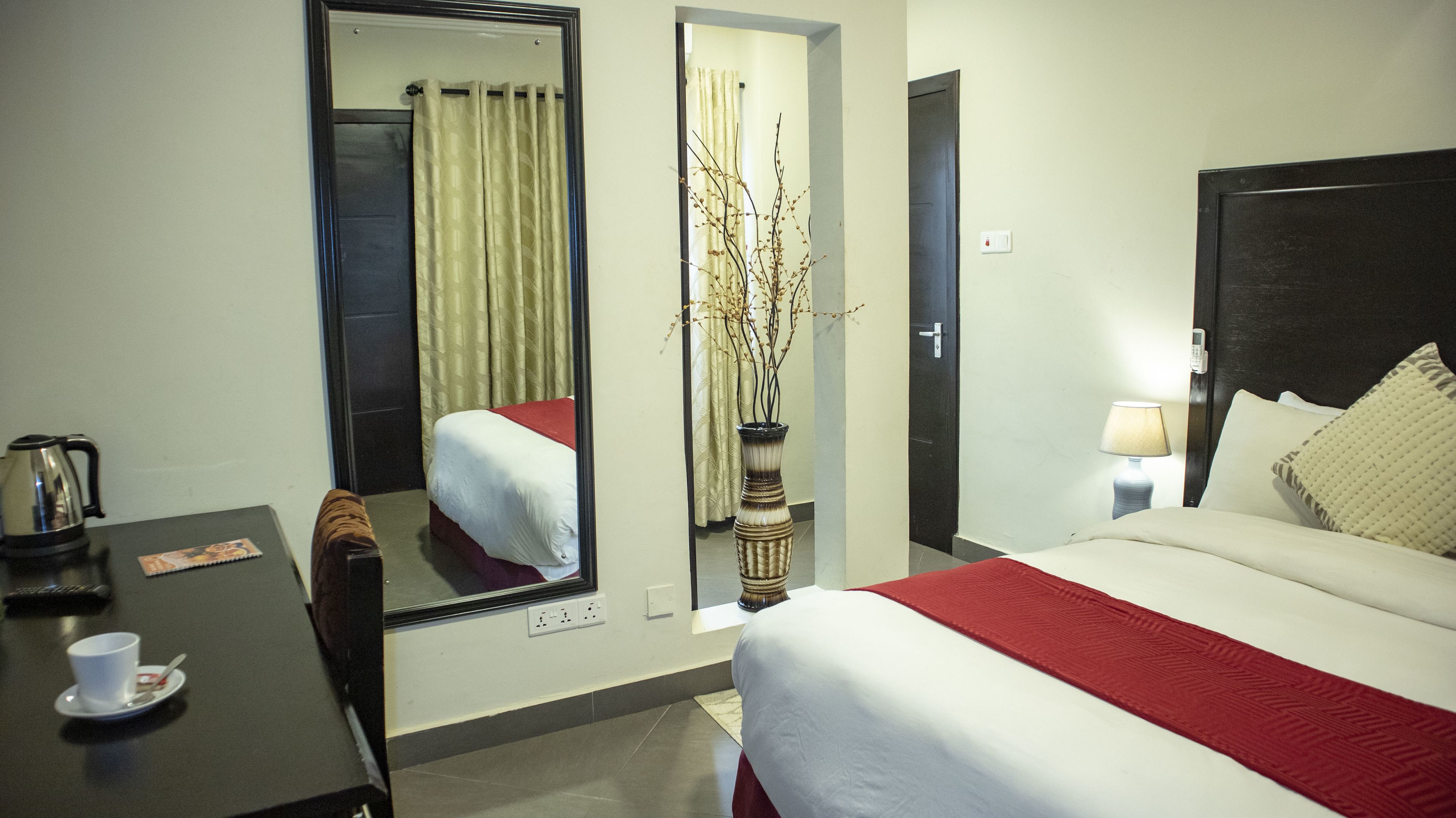 Une chambre d’hôtel avec un lit, un bureau, un miroir, un vase avec des branches, une lampe et un téléphone.