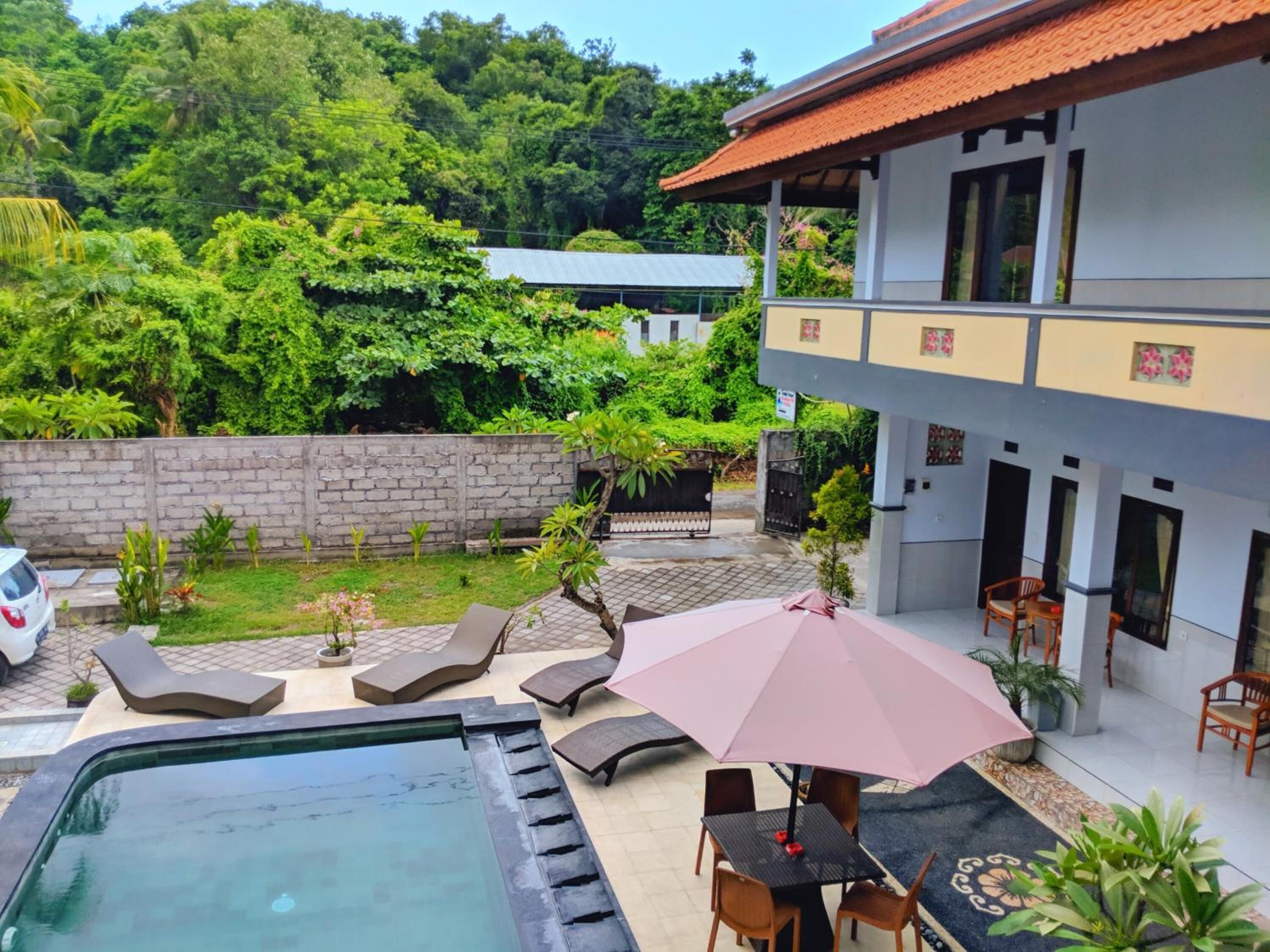 A pool area with lounge chairs and a table under a pink umbrella, surrounded by a garden and a building with a tiled roof.