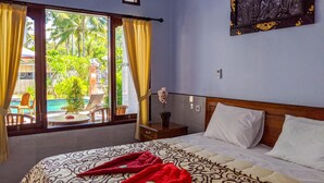 A hotel room with a bed, a nightstand, a window with curtains, and a view of a pool and greenery outside. - Serangan Inn Mimba (Padangbai)