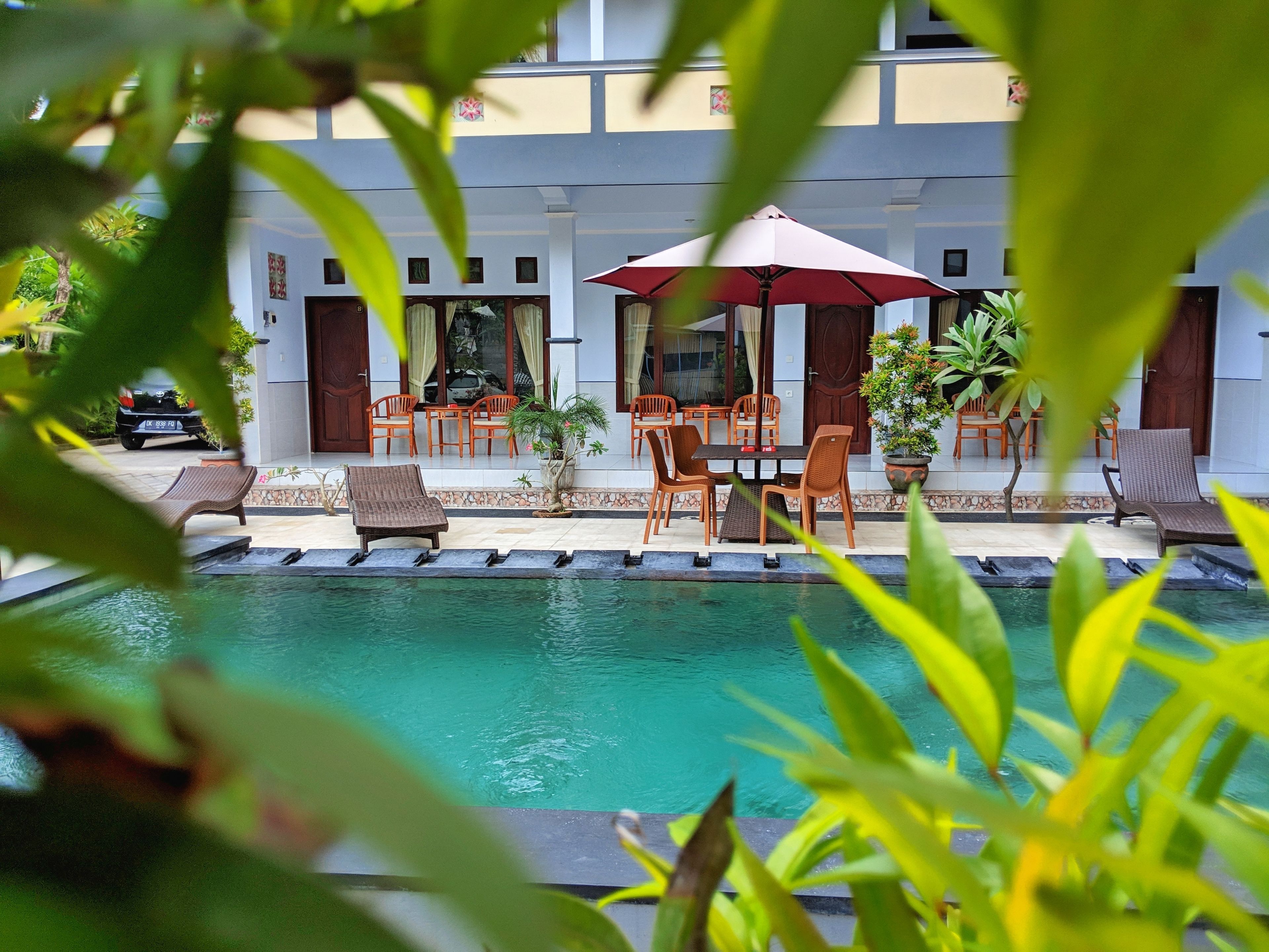 A pool area with lounge chairs and a table, surrounded by potted plants and a building with multiple windows and doors.