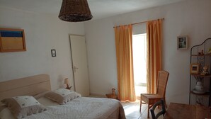 A bedroom with a bed, a chair, a table, a wicker lamp, and a window with curtains. - Ferme Le Grand Cordy (Grignan)