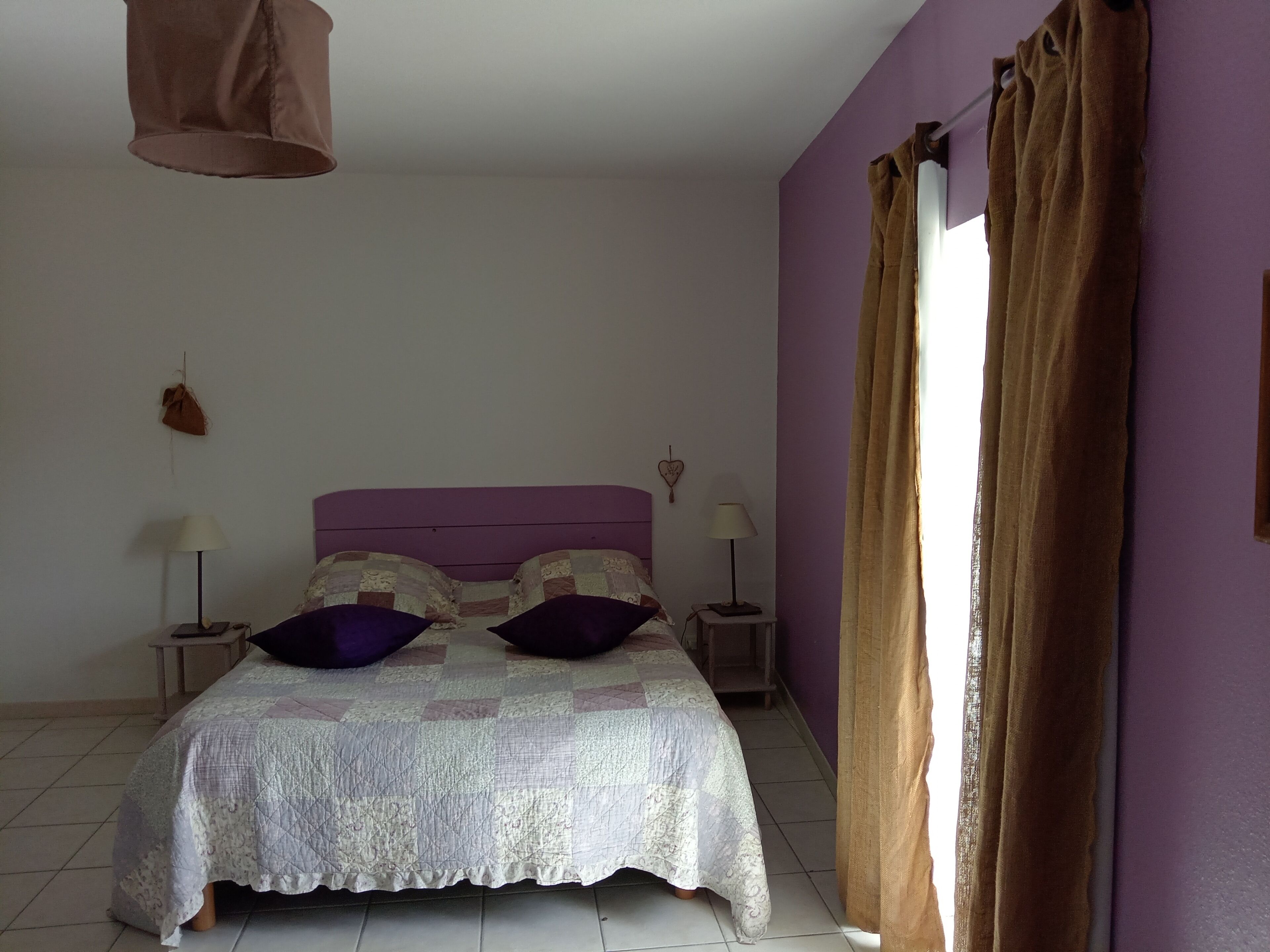 A bedroom with a bed, two bedside lamps, a hanging light fixture, a heart-shaped decoration, and a window with curtains.