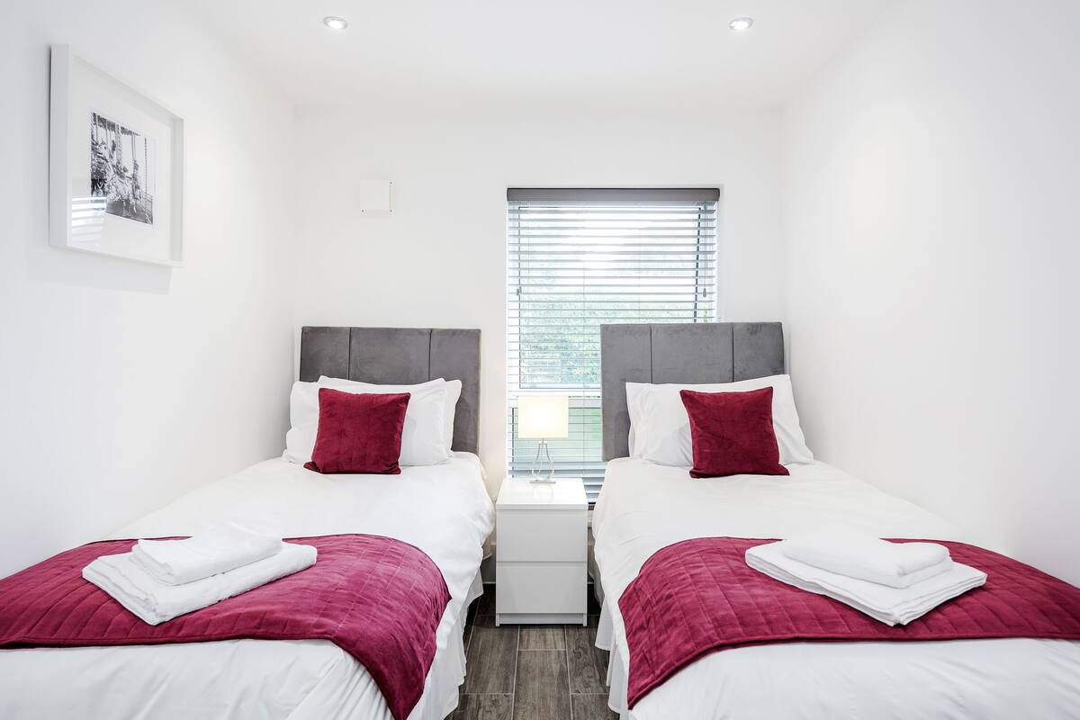 A hotel room with two beds, each with a red and white bedspread, white pillows, and a red throw blanket. There is a nightstand between the beds with a lamp, and a framed picture on the wall.