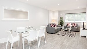 Un salon moderne avec une table à manger en verre, des chaises blanches, des canapés gris et une grande fenêtre avec des stores.