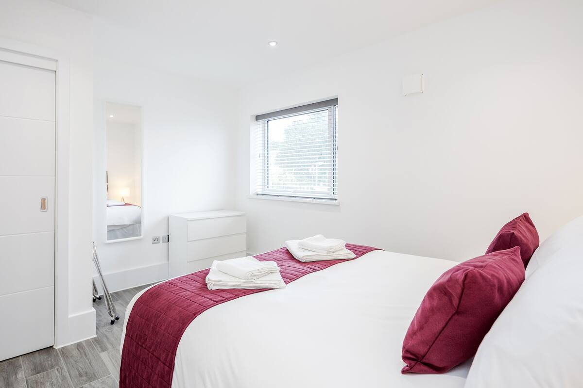 A modern bedroom with a neatly made bed, a window with blinds, a bedside table, and a mirror.
