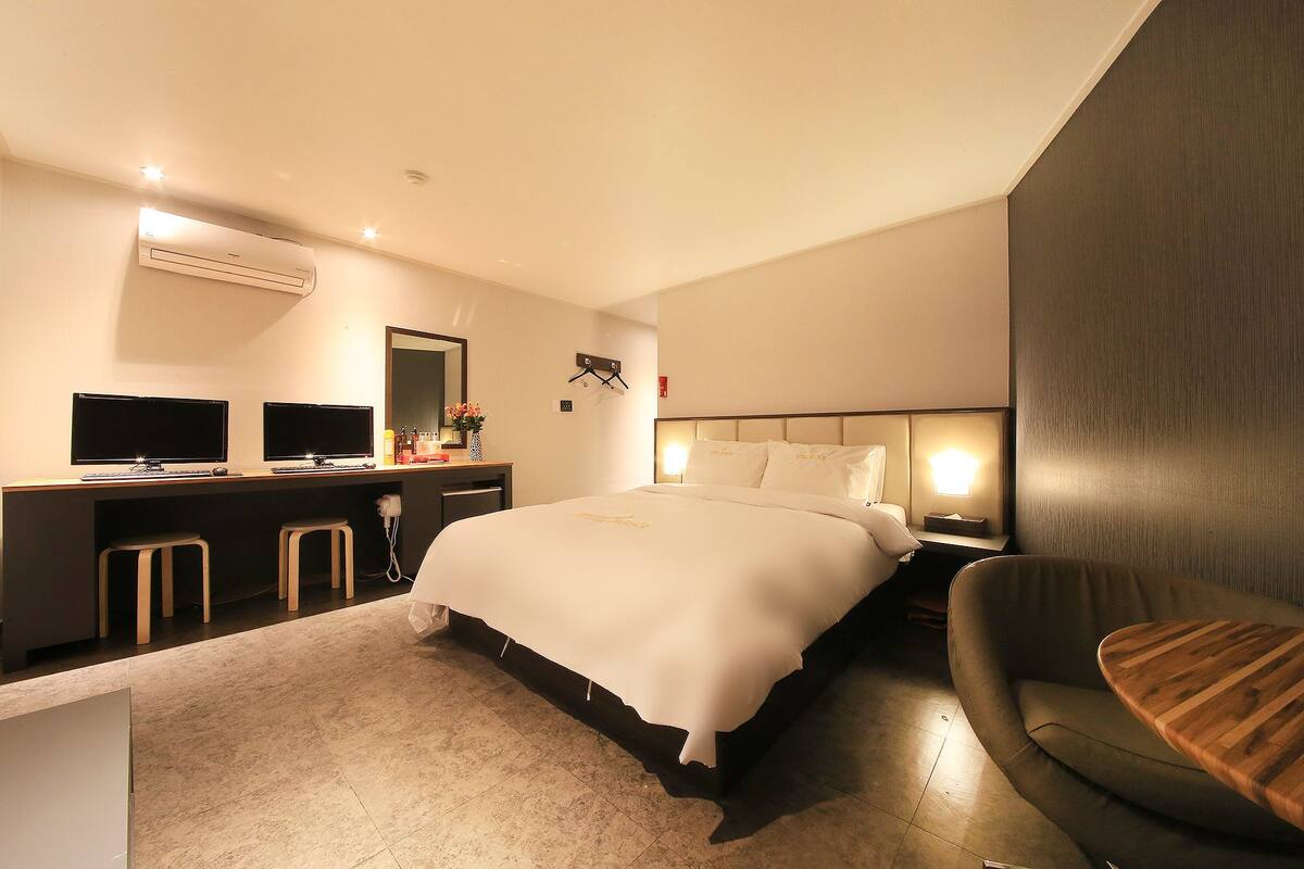 A modern hotel room with a large bed, a desk with two monitors, a chair, and a small table.