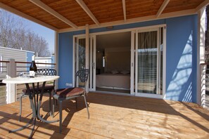 Un espacio exterior cubierto con una mesa y sillas, una puerta corrediza de vidrio que da a un dormitorio y una pared exterior azul.