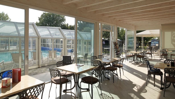 Un espacio interior amplio con ventanas grandes que dan a una piscina y follaje. Mesas y sillas están dispuestas para comer o descansar.