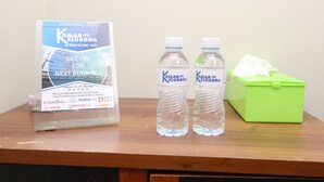 Ein Willkommensschild für Kamar Keluarang, zwei Wasserflaschen und ein grüner Mülleimer auf einer Holzfläche.