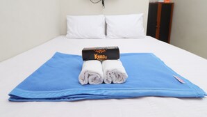 Ein ordentlich gemachtes Bett mit einem blauen Handtuch und einem aufgerollten weißen Handtuch auf einer blauen Matte, eine schwarze Box auf der Matte, ein an der Wand befestigtes Fernsehgerät und ein Holzkabinent mit Flaschen.
