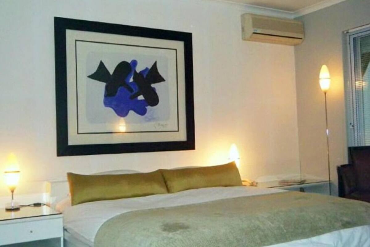 Habitació d'hotel amb llit, tauletes de nit, una obra d'art abstracta emmarcada, un aparell de climatització i una làmpada de peu.