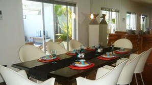 A dining area with a long table set for six, white chairs, and a view of the outdoors through large windows. - Maison Fontainbleau (Cape Town)