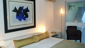 A hotel room with a bed, a framed abstract artwork, an air conditioning unit, a bedside table, a lamp, a chair, and a window with blinds. - Maison Fontainbleau (Cape Town)