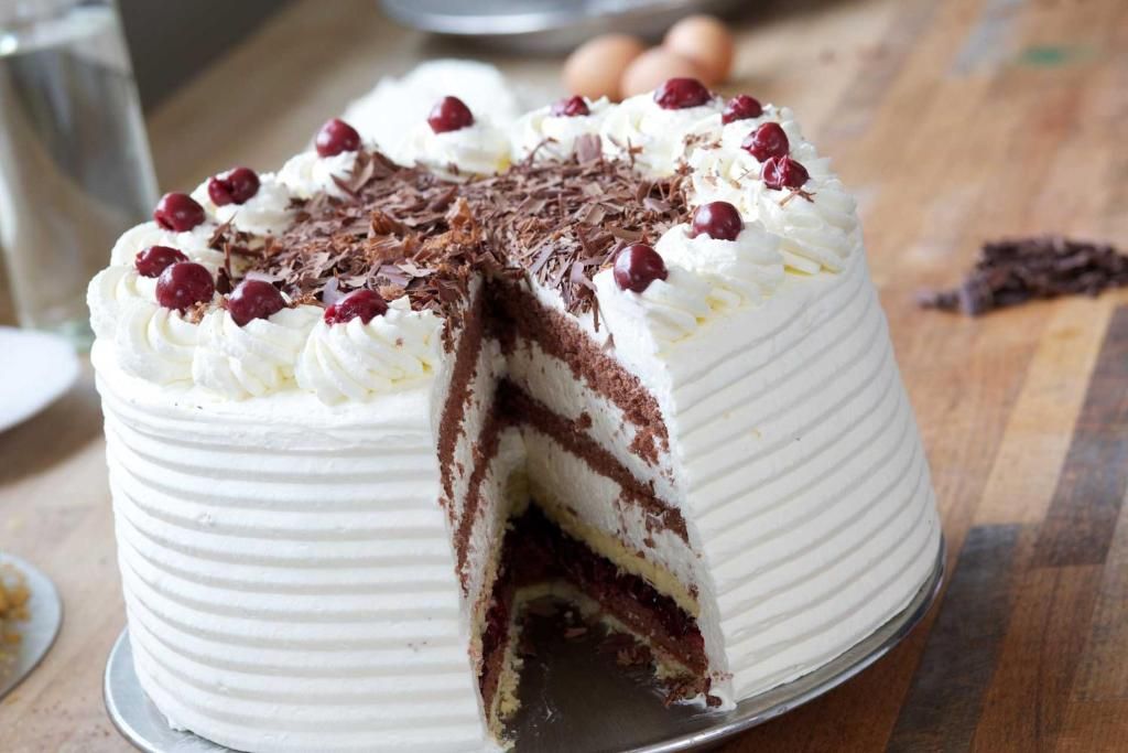 A cake with white frosting and chocolate layers, garnished with cherries and chocolate shavings.
