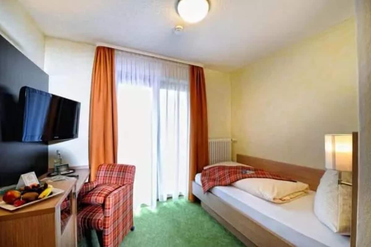 A hotel room with a bed, a desk with a television, a chair, and a window with curtains.