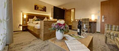 A hotel room with a bed, a TV, a coffee table with a vase of flowers, a magazine, and a plaid armchair.